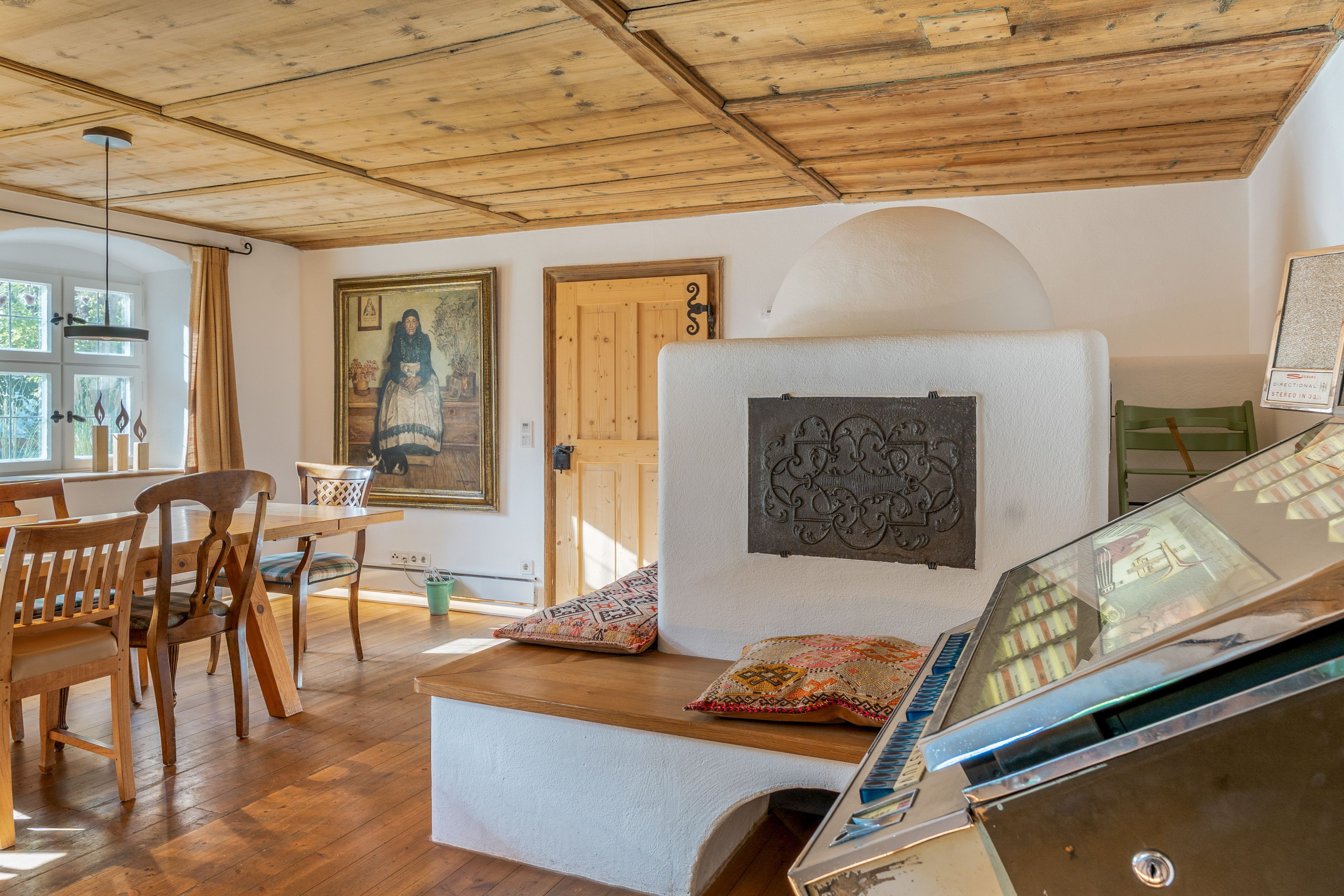 A room with wooden flooring, a dining table with chairs, a sofa with cushions, a wall-mounted TV, and a door with a wooden frame.
