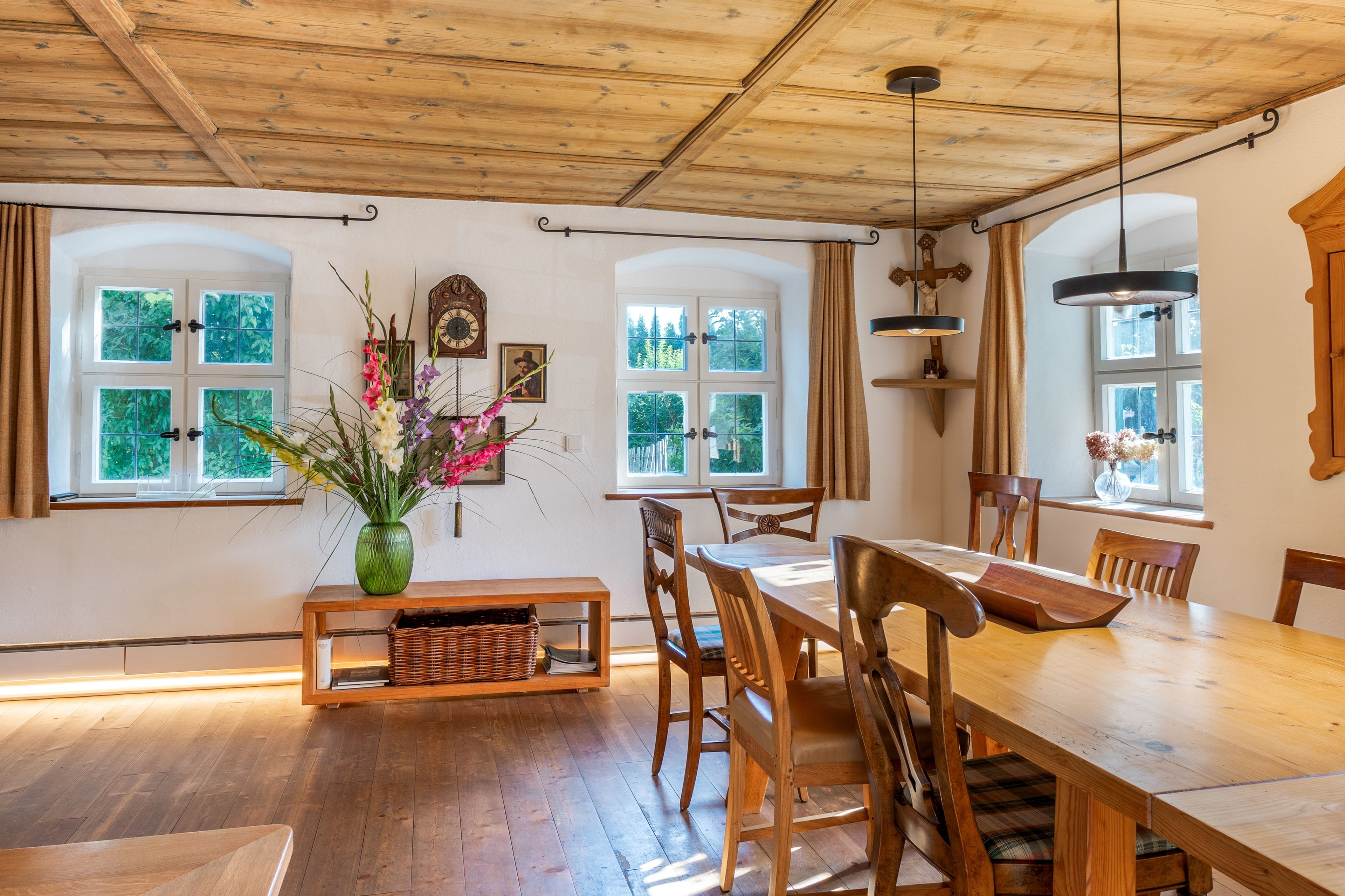 A dining area with wooden furniture, a vase of flowers, and large windows with curtains.
