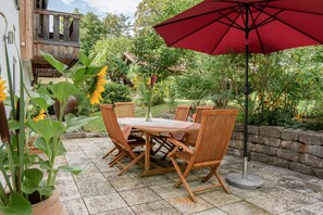 Eine Terrasse im Freien mit Tisch und Stühlen, umgeben von Grünpflanzen und einem Holzzaun.