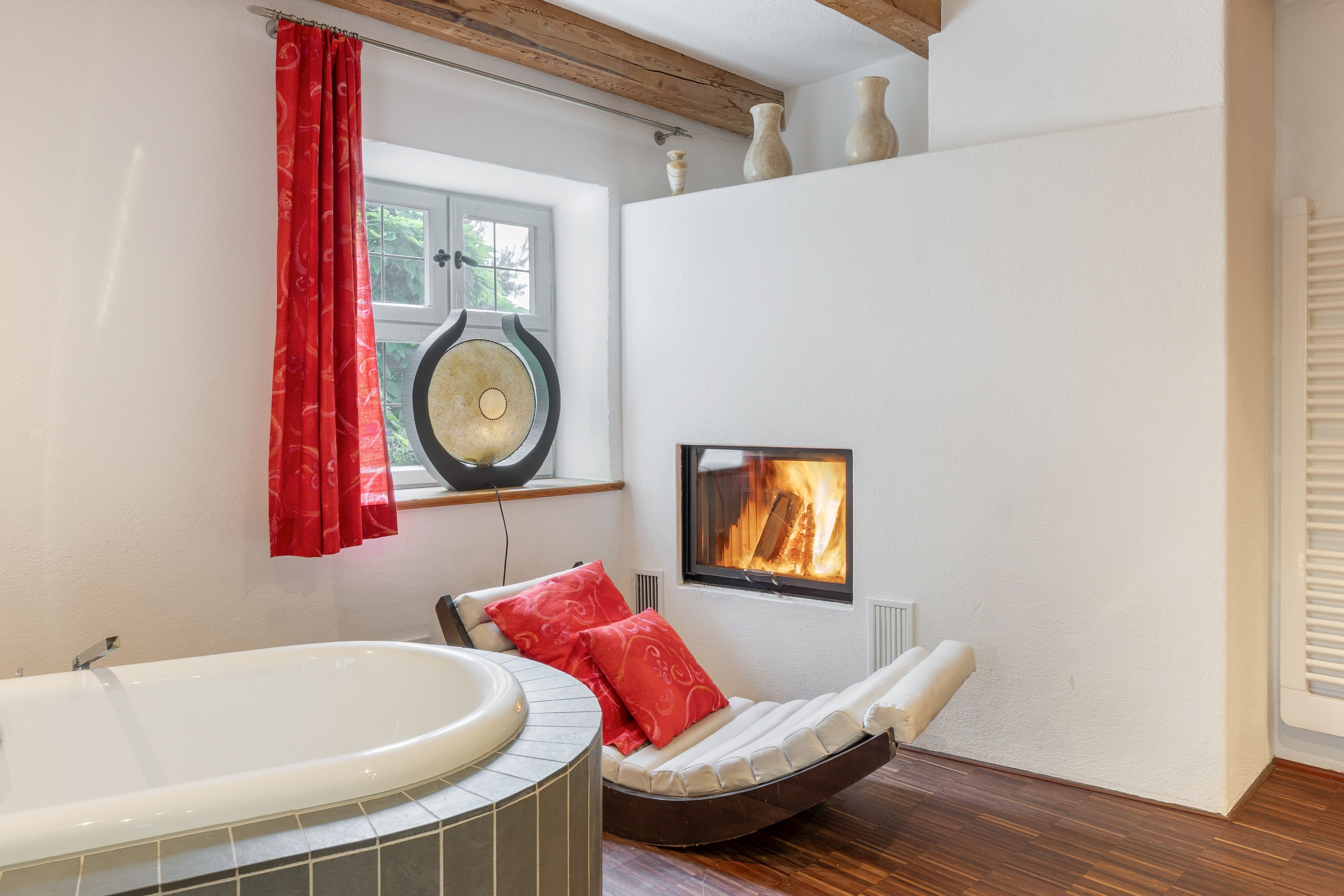 Ein Badezimmer mit einer Badewanne, einem roten Vorhang, einem Fenster, einem Kamin und einem Sessel mit Kissen.