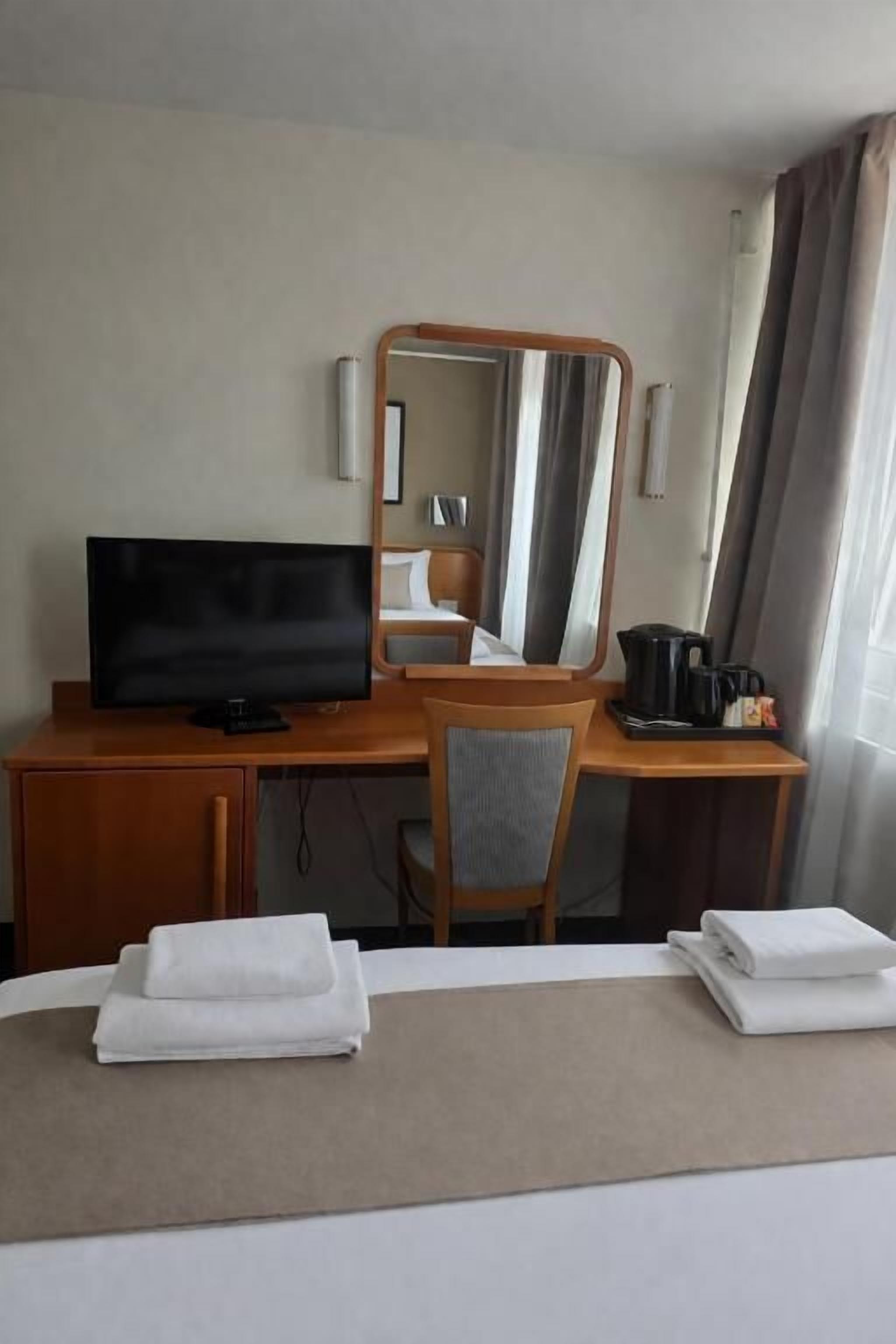 A hotel room with a bed, a desk with a television, a chair, a mirror, and a window with curtains.