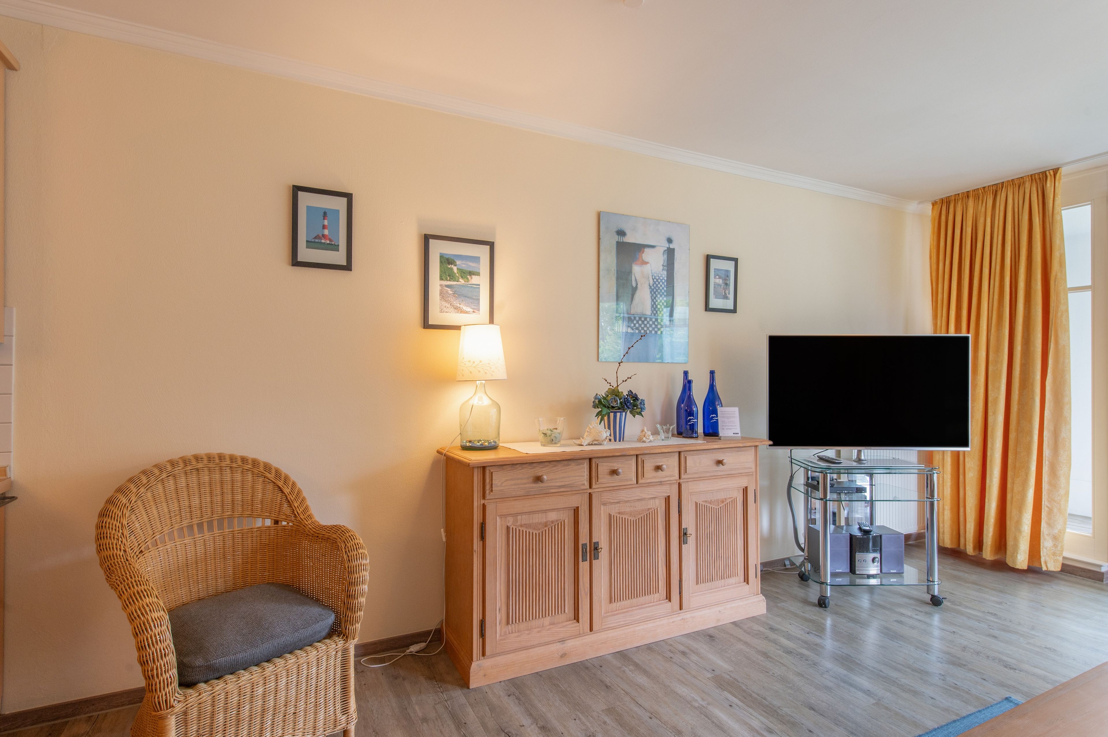 Ein Wohnzimmer mit Fernseher, einem Korbstuhl, einem Holzschrank und Wandbildern.