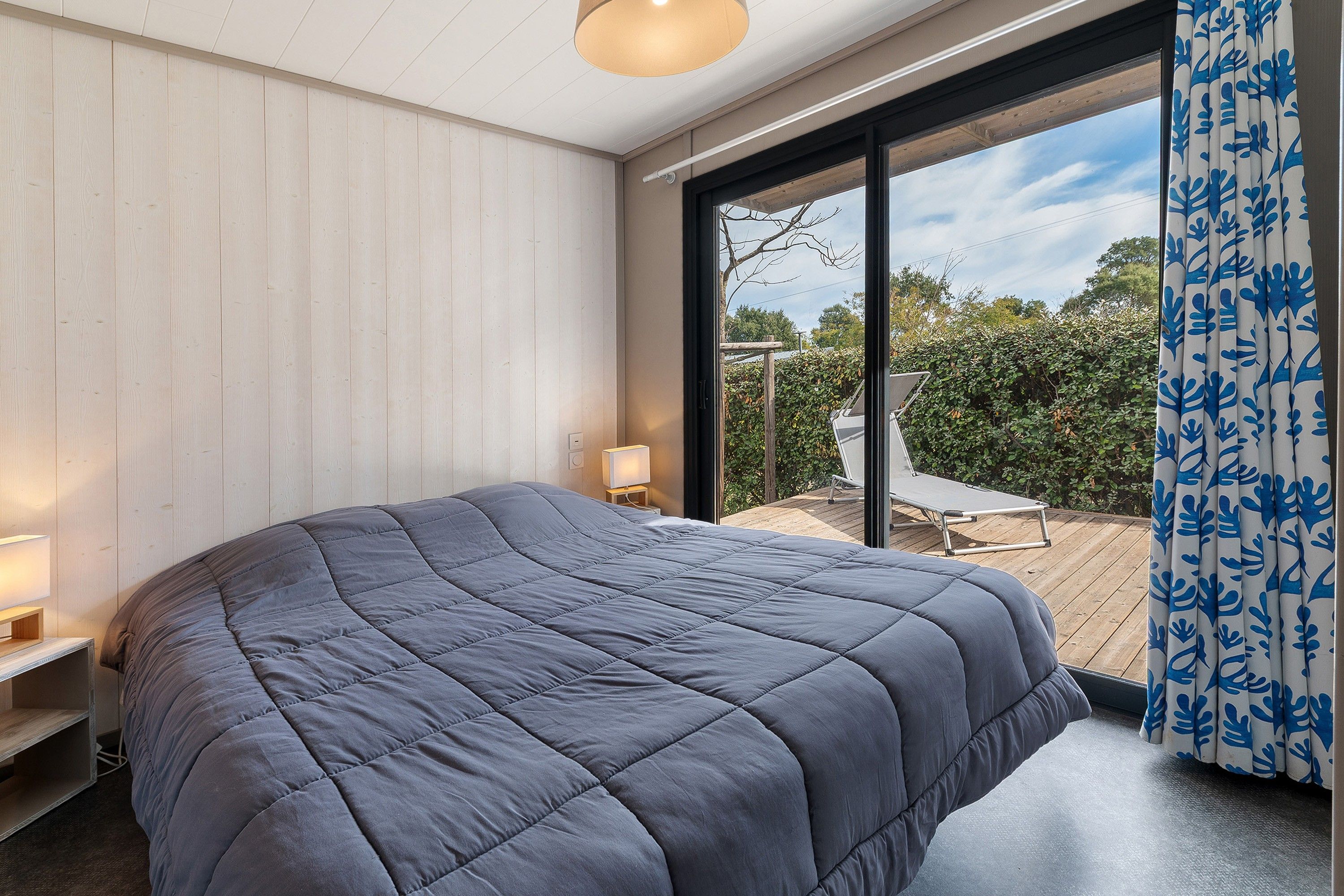 A bedroom with a large bed, a bedside table, a lamp, and a view of a deck and greenery outside.