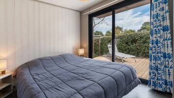 A bedroom with a large bed, a bedside table, a lamp, and a view of a deck and greenery outside.