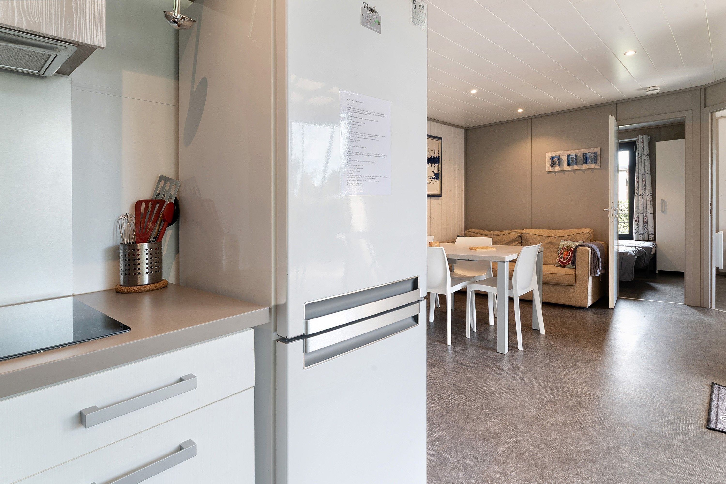 A modern kitchen with a refrigerator, a dining table with chairs, and a living area with a sofa and a window.