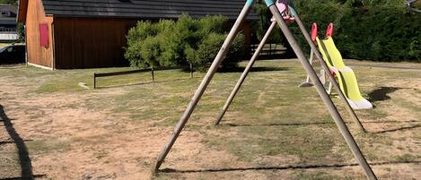 A playground with a slide and swings, a wooden barn, and a hilly landscape in the background.