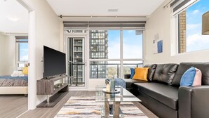A modern living room with a sofa, a flat-screen TV, a glass coffee table, and a large window with a city view. - Modern High Rise Condo w Private Patio Downtown (Kitchener)
