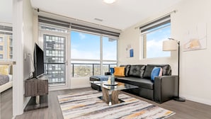 A modern living room with a sofa, a coffee table, a TV, and large windows. - Modern High Rise Condo w Private Patio Downtown (Kitchener)