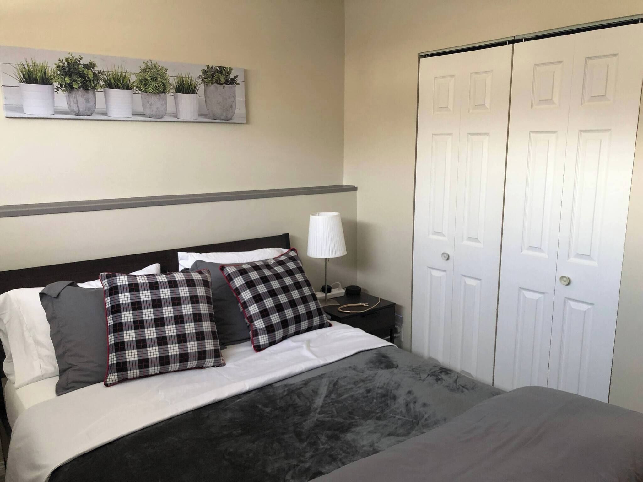 A neatly made bed with a grey blanket, two black and white checkered pillows, a bedside lamp, and a wall-mounted plant shelf.