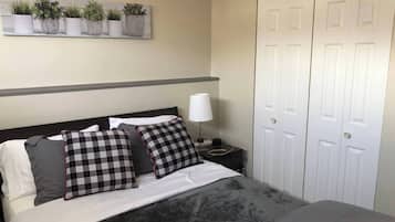 A neatly made bed with a grey blanket, two black and white checkered pillows, a bedside lamp, and a wall-mounted plant shelf.