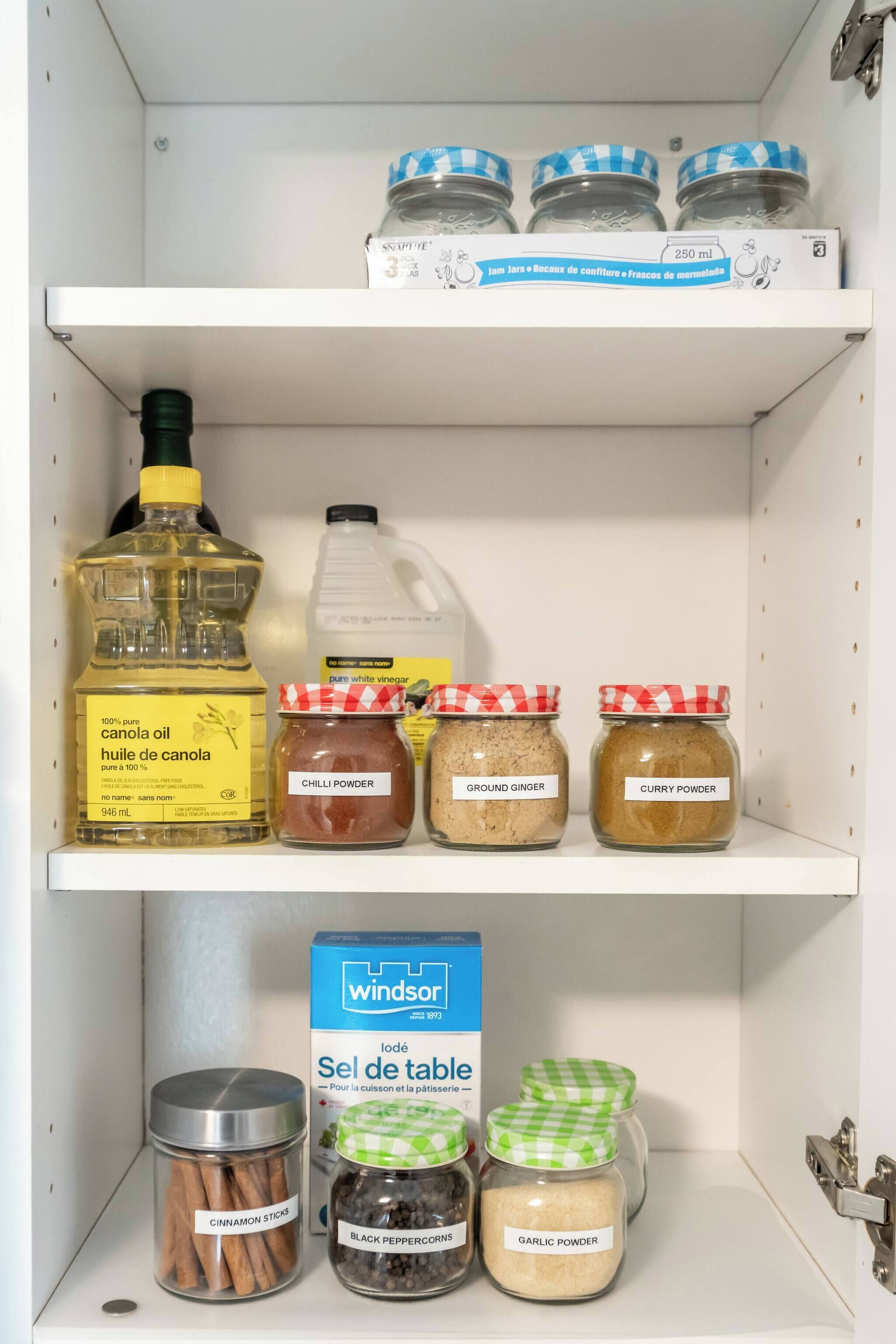 A pantry shelf with various food items including canola oil, vinegar, chili powder, ground ginger, curry powder, cinnamon sticks, black peppercorns, garlic powder, and a box of Windsor table salt.