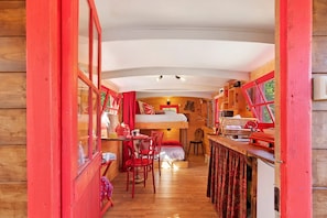 Un étroit couloir en bois, avec des fenêtres à cadre rouge, un lit superposé, une table à manger avec des chaises, et un coin cuisine doté de placards en bois et d’un évier.