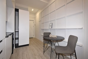 A modern kitchen with a round table and two chairs, a white wall with a shelf, and a door leading to another room. - Modern Studio Apartment in the City Centre (Yerevan)