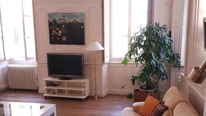 Een woonkamer met een bank, een salontafel, een televisie, een schilderij, een lamp en een grote potplant.