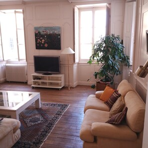 Ein Wohnzimmer mit einer Couch, einem Couchtisch, einem Fernseher, einem Gemälde, einer Lampe und einer großen Pflanze.