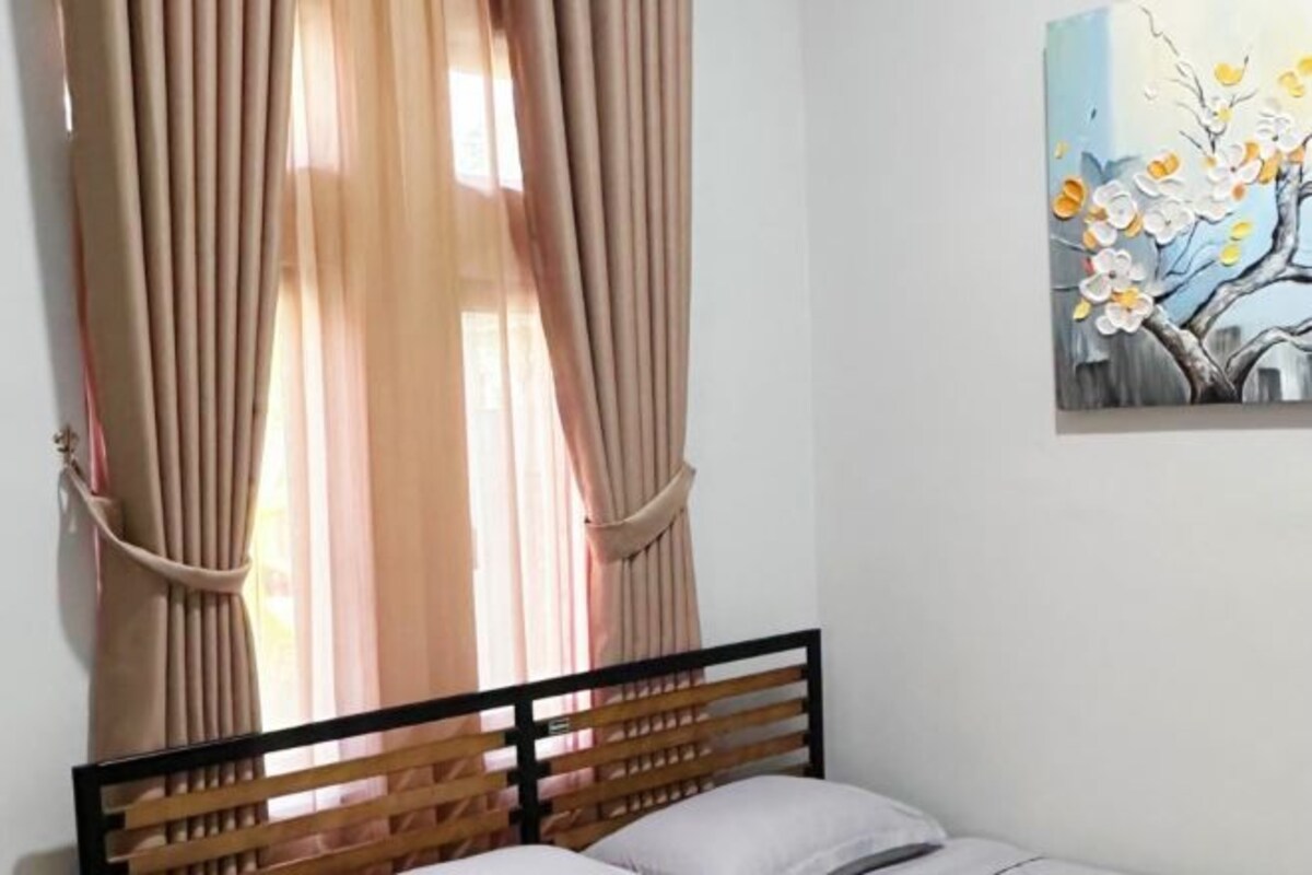 Un dormitorio con cabecera de madera, cortinas color beige y un cuadro de flores en la pared.