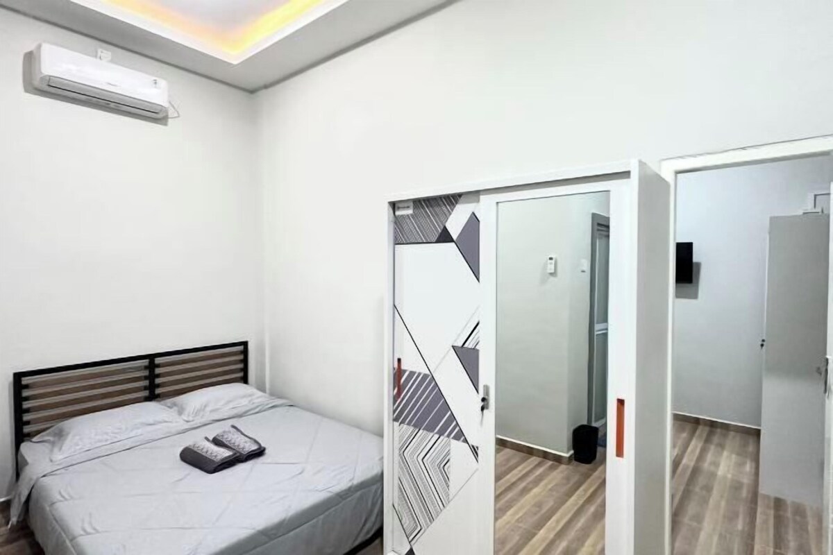 A bedroom with a bed, an air conditioner, a geometric-patterned door, and a hallway with a wooden floor.