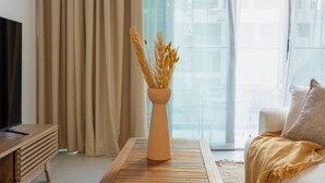 A modern living room with a flat-screen TV, a wooden coffee table, a vase with dried plants, a sofa, and a large window with curtains. - Bloomfields Studio in Park View   (Abu Dhabi)