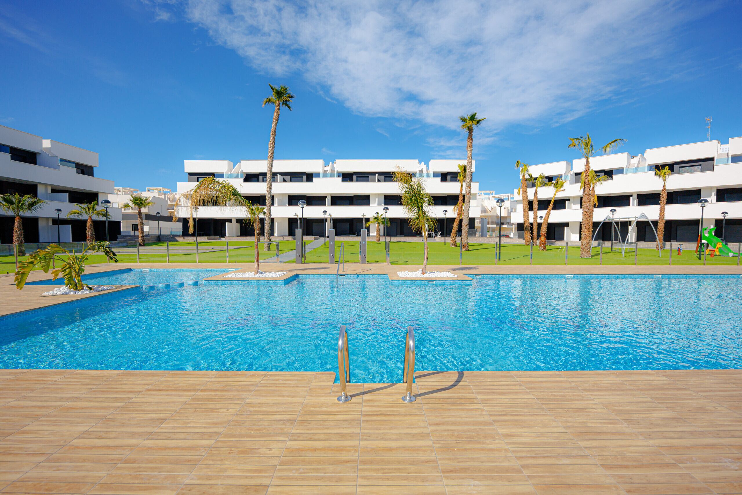 Une piscine dotée d’une terrasse carrelée, entourée de palmiers et de bâtiments modernes.