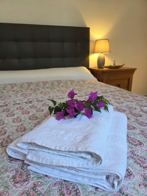Un lit bien fait, recouvert d’une couvre-lit à motifs floraux, une pile de serviettes blanches pliées et un arrangement décoratif de fleurs violettes posé sur le lit.