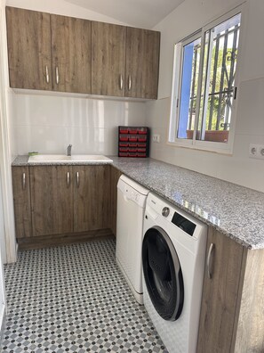 A compact kitchen with a washing machine, wooden cabinets, a tiled floor, and a window with a view. - Charming Guest House with a Private Pool near Gandia (Marxuquera)