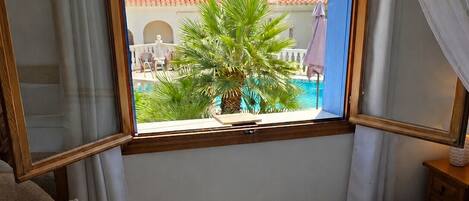 Quarto de hotel com cama, janela com vista para uma piscina e palmeira, e uma cadeira.