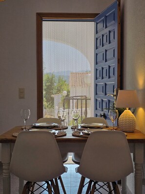 Une table à manger dressée avec des assiettes, des verres et des couverts, une lampe, et une vue par une fenêtre ornée d’une porte à motifs.