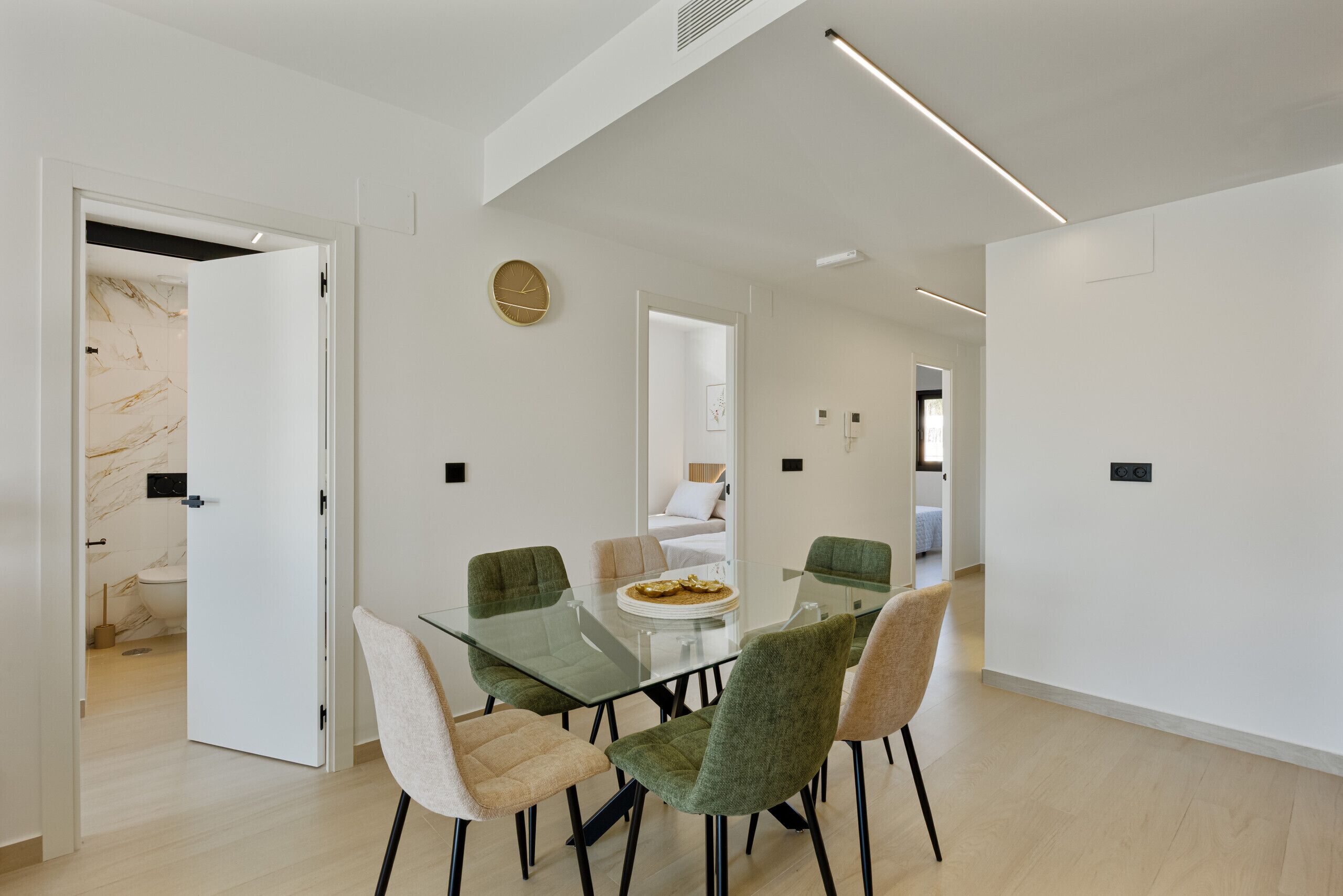 Un comedor moderno con una mesa y sillas de vidrio, una puerta blanca que da al baño y una lámpara en el techo.