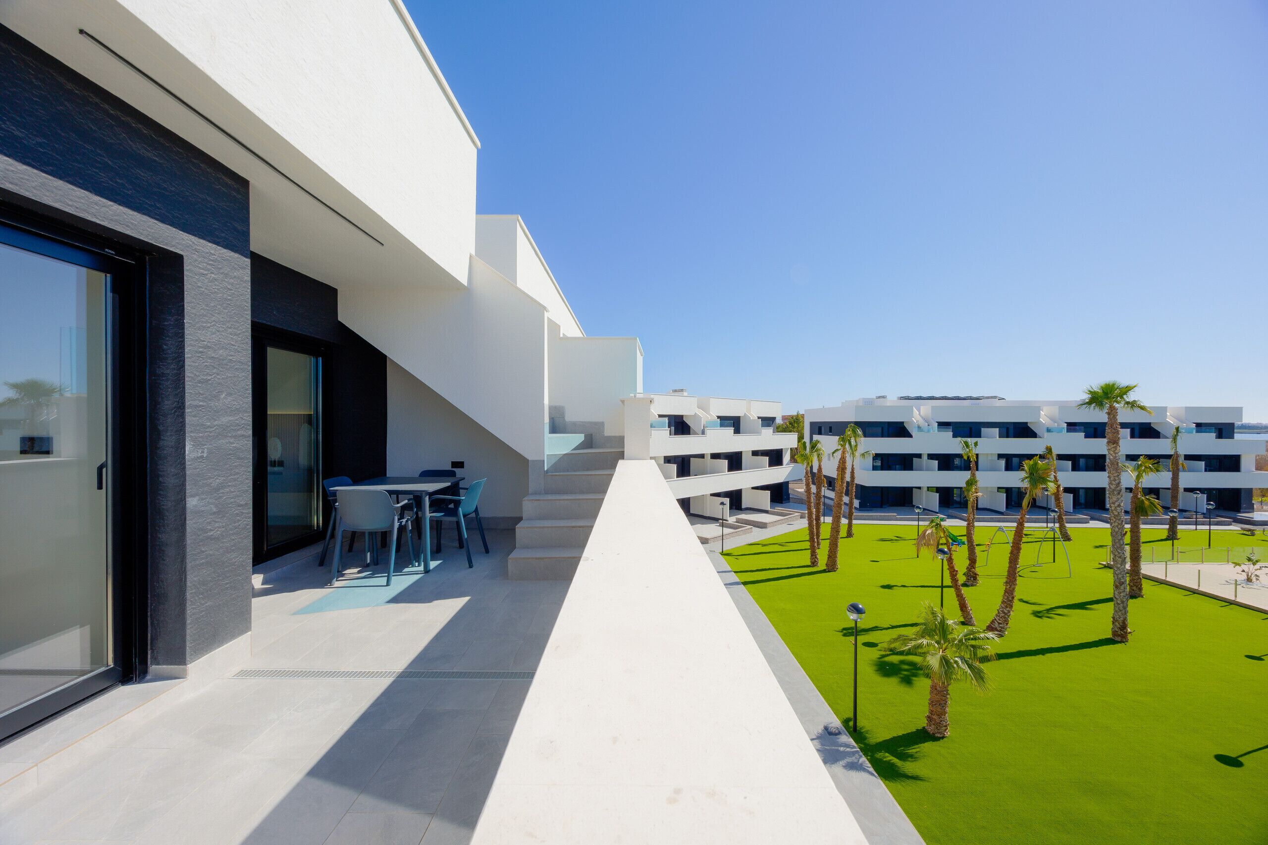 Un edificio moderno con balcón, mobiliario exterior y un césped bien cuidado con palmeras.