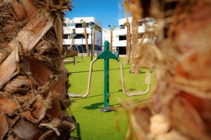 Ein Spielplatz mit grünen Trainingsgeräten, umgeben von Palmen und einem Gebäude im Hintergrund.