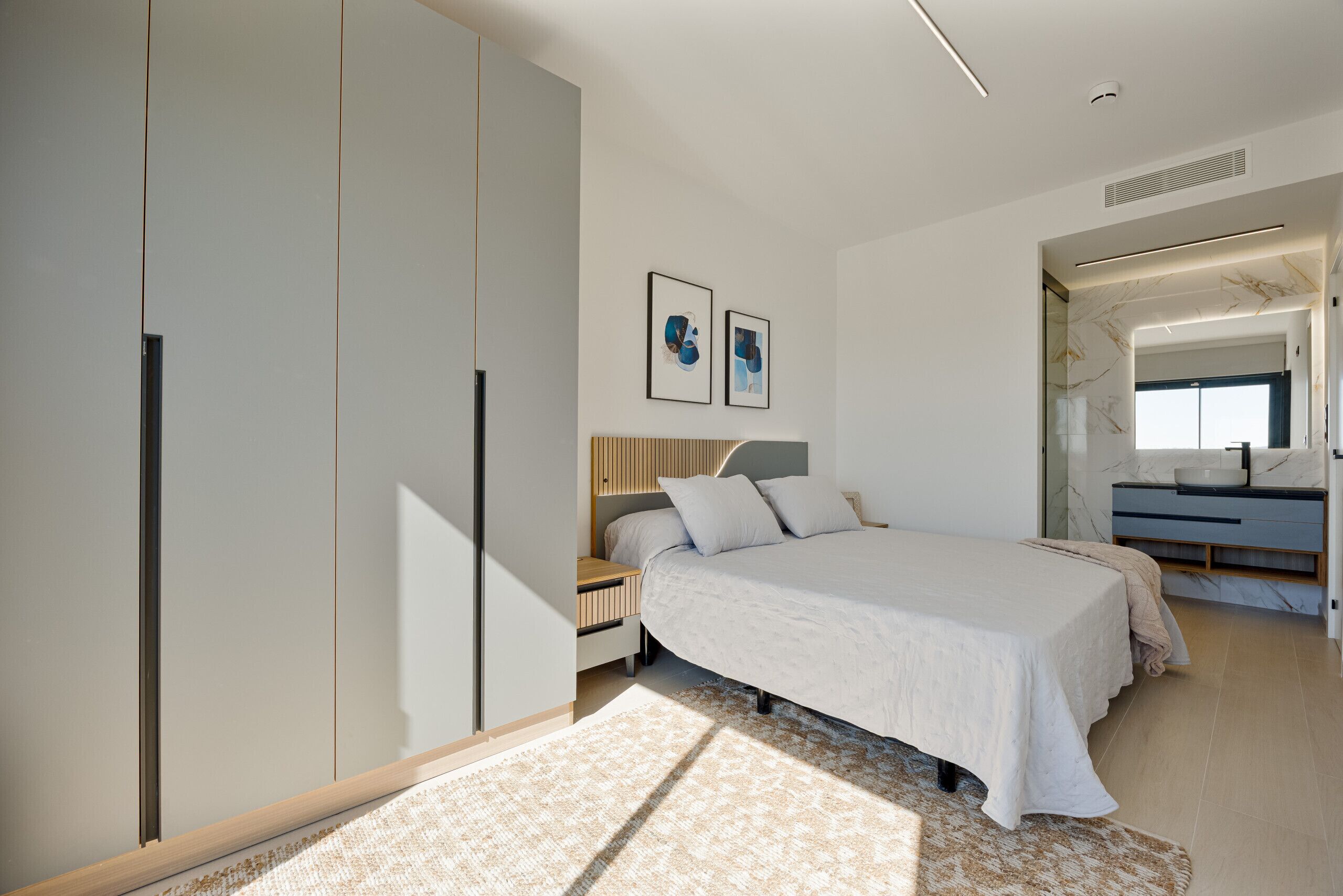 A modern bedroom with a large bed, a wardrobe, a bedside table, and a bathroom with a mirror and sink.