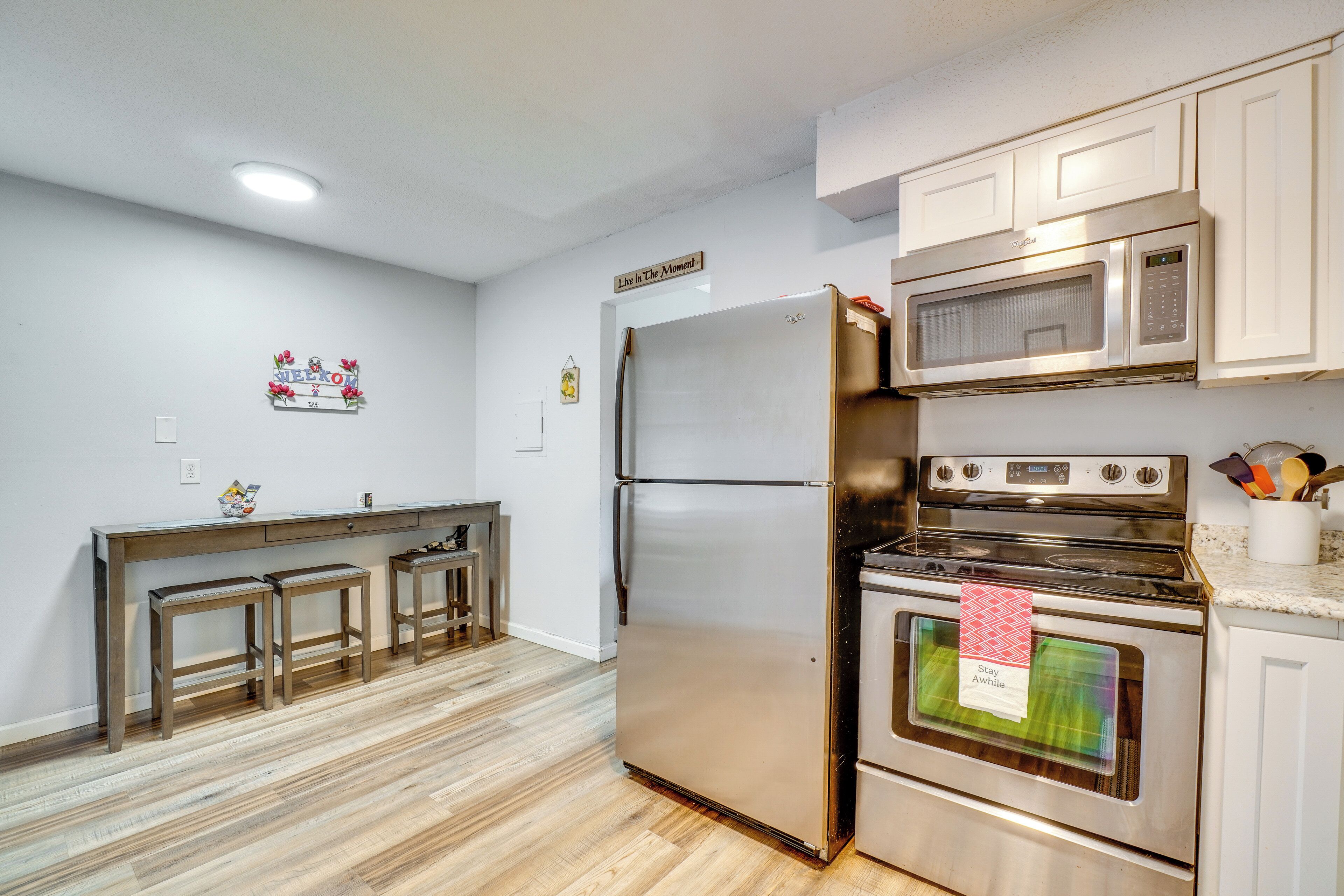 Una cocina con refrigerador de acero inoxidable, horno, microondas, taburetes de bar de madera y un letrero que dice 'Vive el momento'.