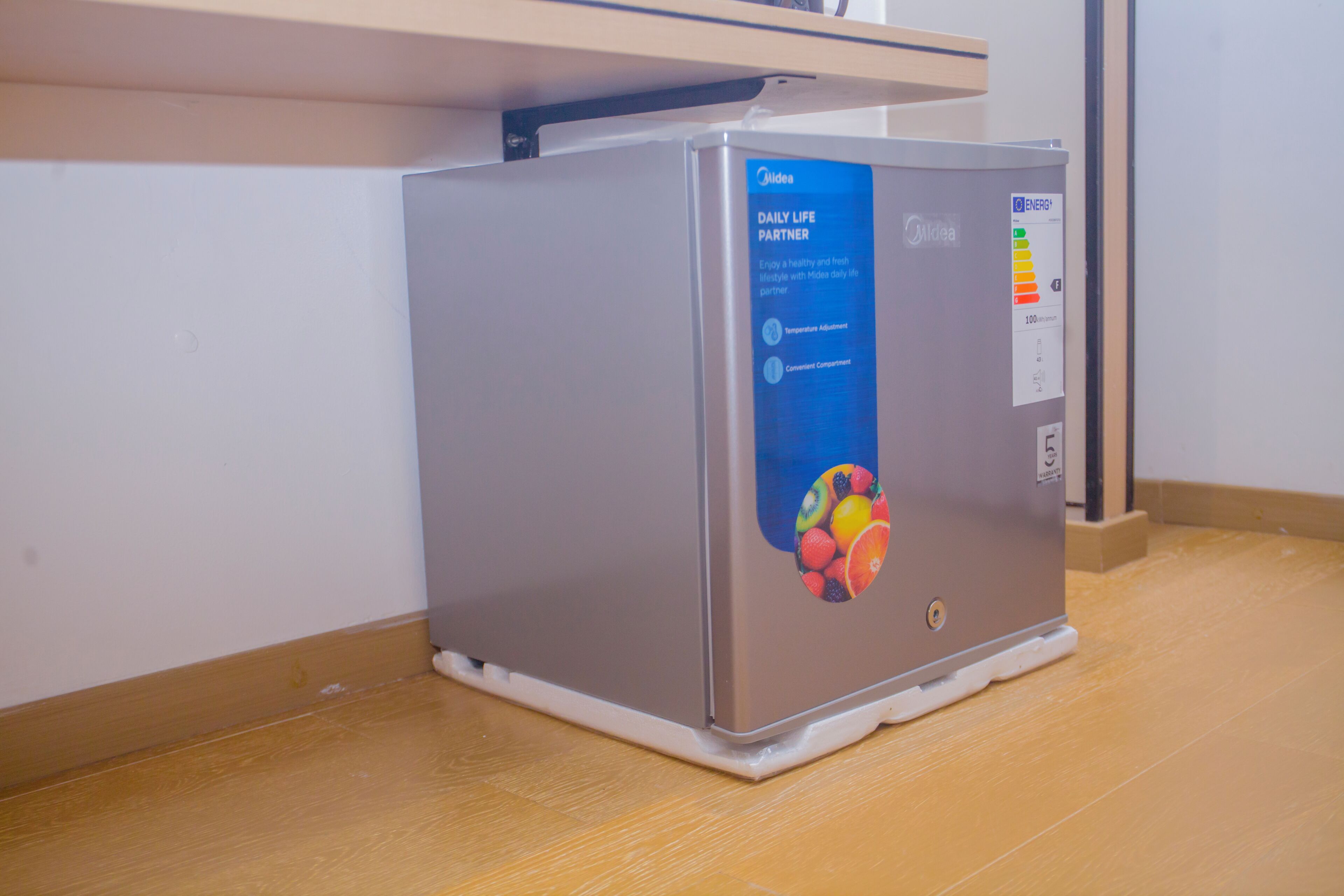 A compact, silver-colored refrigerator with a blue label on the front, placed under a wooden shelf in a room with wooden flooring.