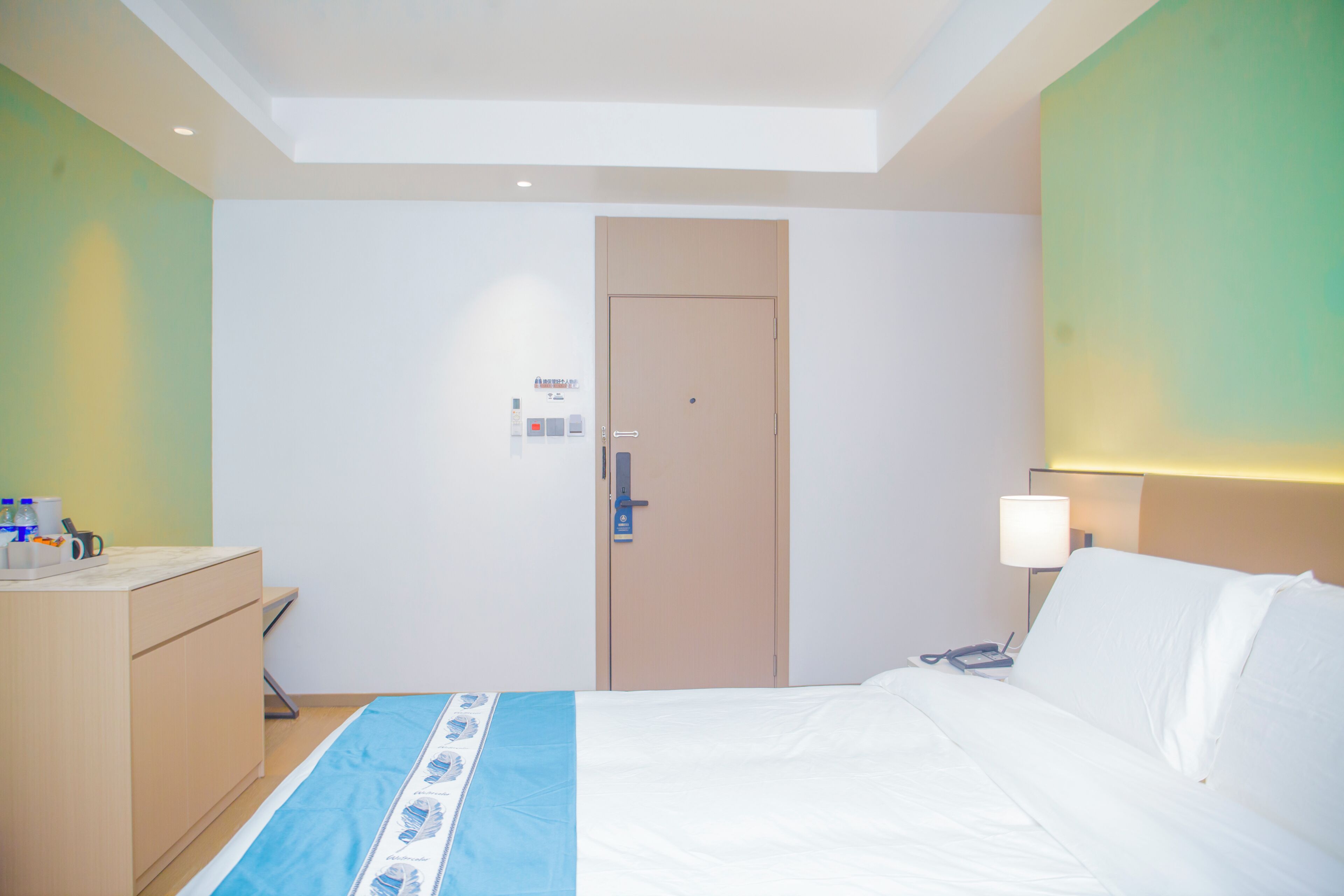 Una habitación de hotel con cama, mesita de noche, lámpara y una puerta con panel de control.