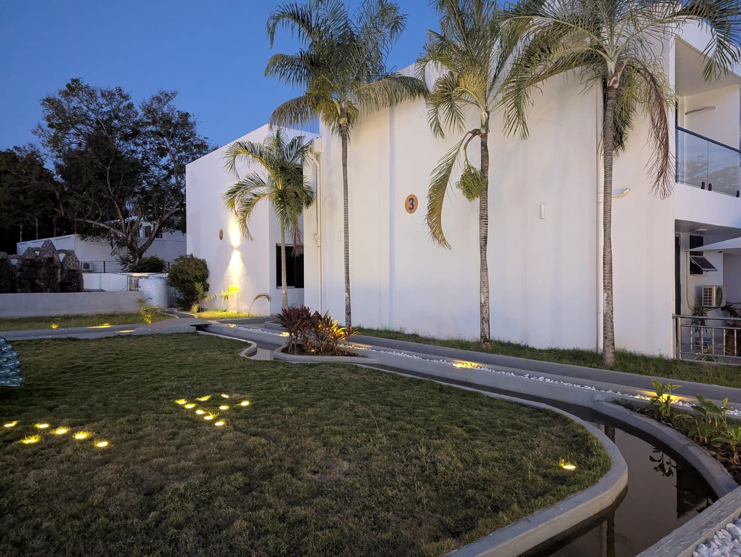 Un edificio moderno con fachada blanca, palmeras y un césped cuidado con luces decorativas.