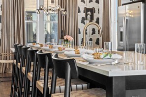 Ein moderner Essbereich mit einem langen Tisch, gedeckt für eine Mahlzeit, schwarzen Stühlen, einer zentralen Kochinsel mit Spüle und einem großen Kühlschrank.