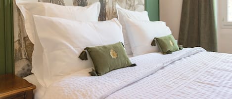 Una cama bien hecha con ropa blanca y dos almohadas verdes, una mesita de noche de madera y una pared con diseño de árbol.