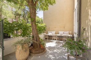 A sunlit outdoor seating area with a table and chairs, surrounded by potted plants and a large tree.