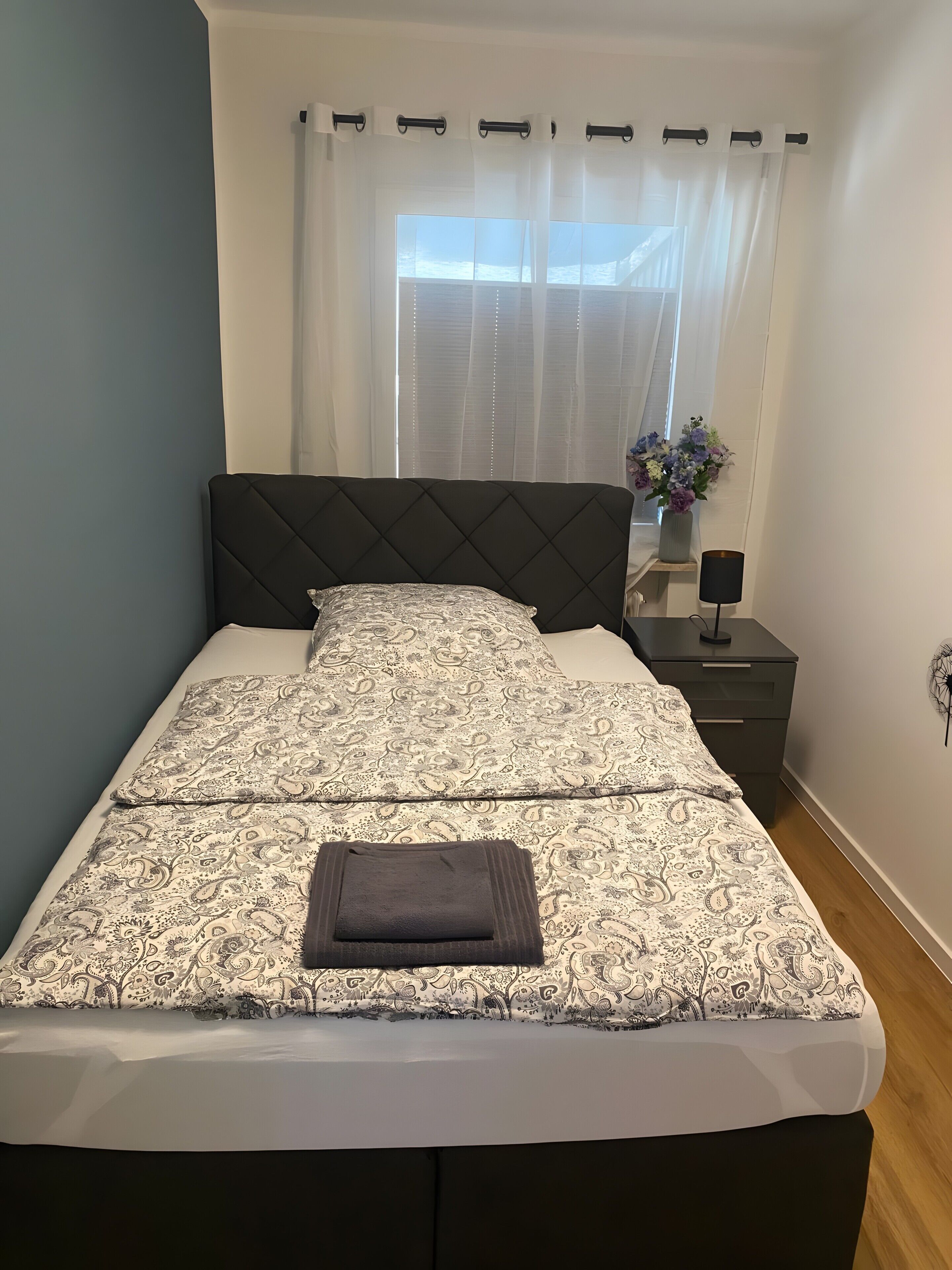 Een bed met een beddengoed met een patroon, een donker hoofdeinde, en een nachtkastje met een lamp.