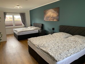 A bedroom with two beds, a window with curtains, a bedside table with a lamp, and a wall-mounted painting.