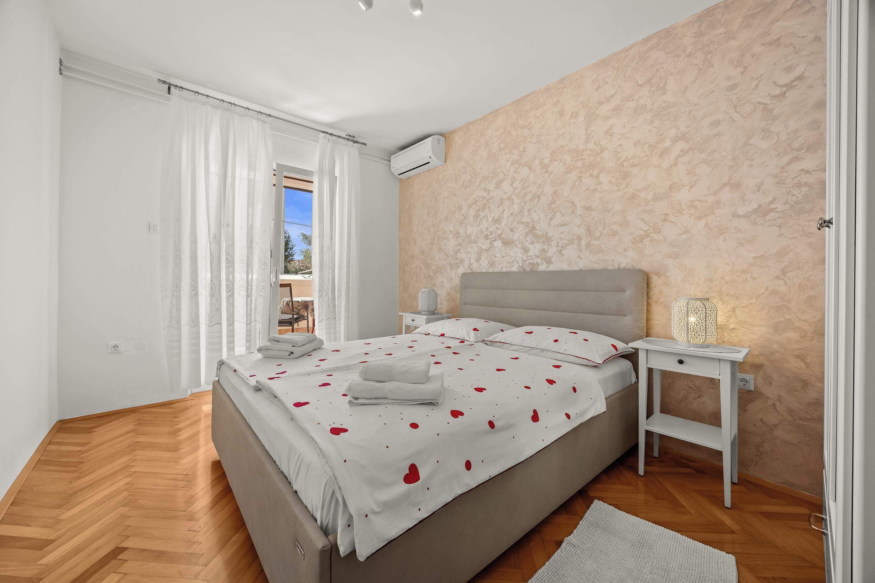 Um quarto com uma cama, uma mesinha de cabeceira com abajur, uma janela com cortinas e piso de madeira.