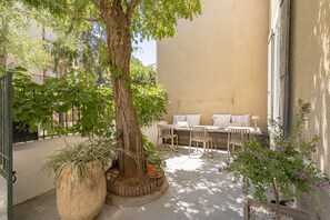 A sunlit outdoor seating area with a table and chairs, surrounded by potted plants and a large tree.
