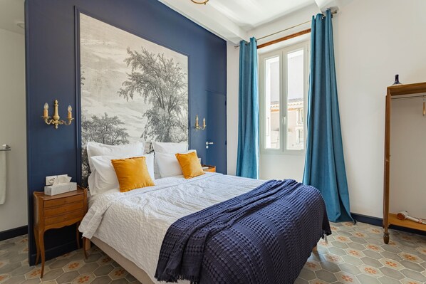 A bedroom with a large bed, a nightstand, a dresser, a window with curtains, and a wall with a large painting.