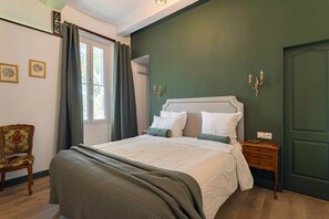 A bedroom with a bed, a chair, a nightstand, a window with curtains, and wall-mounted lighting.