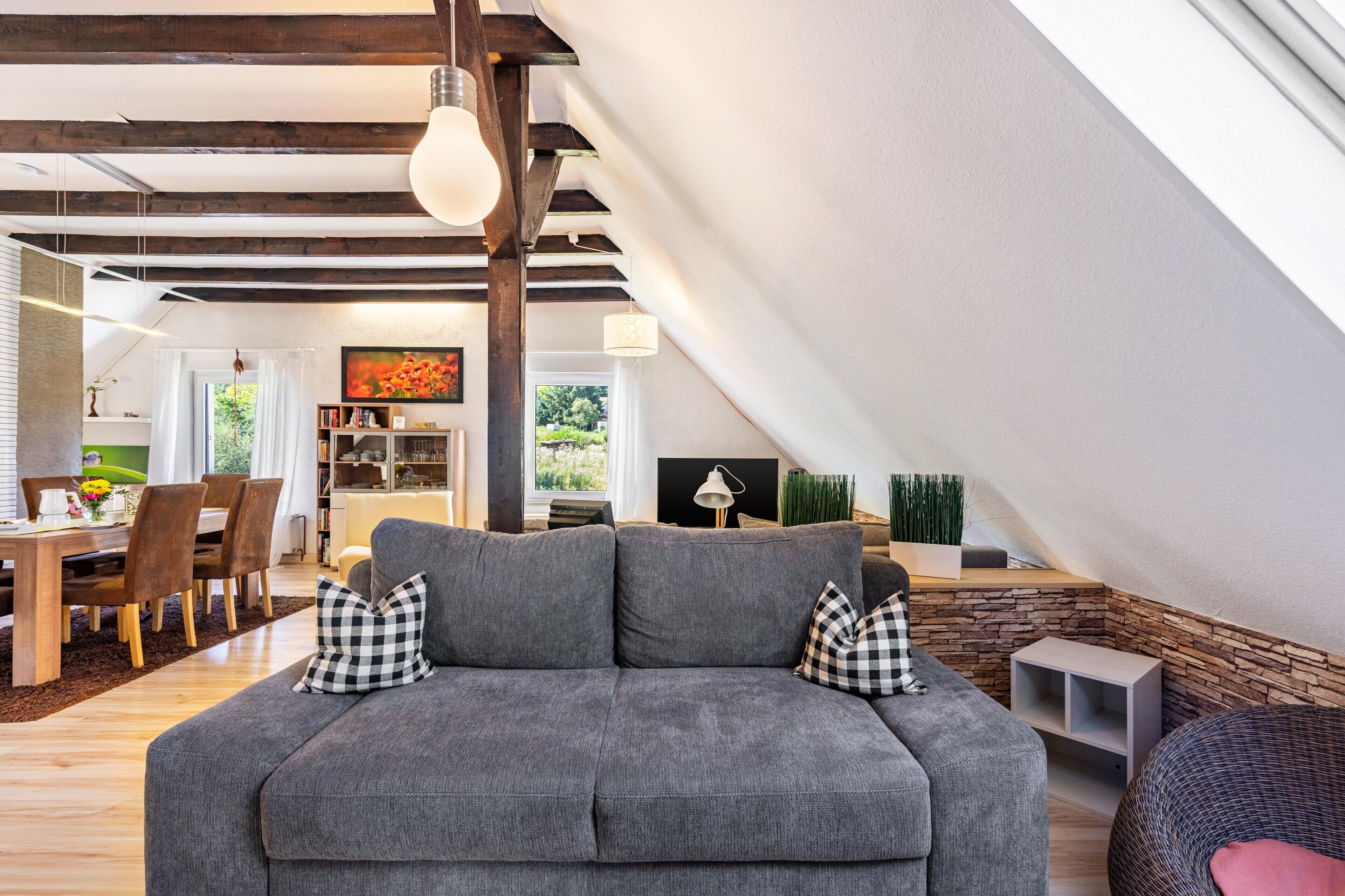 Ein modernes Wohnzimmer mit grauem Sofa, karierten Kissen und einem Essbereich mit Holzmöbeln.