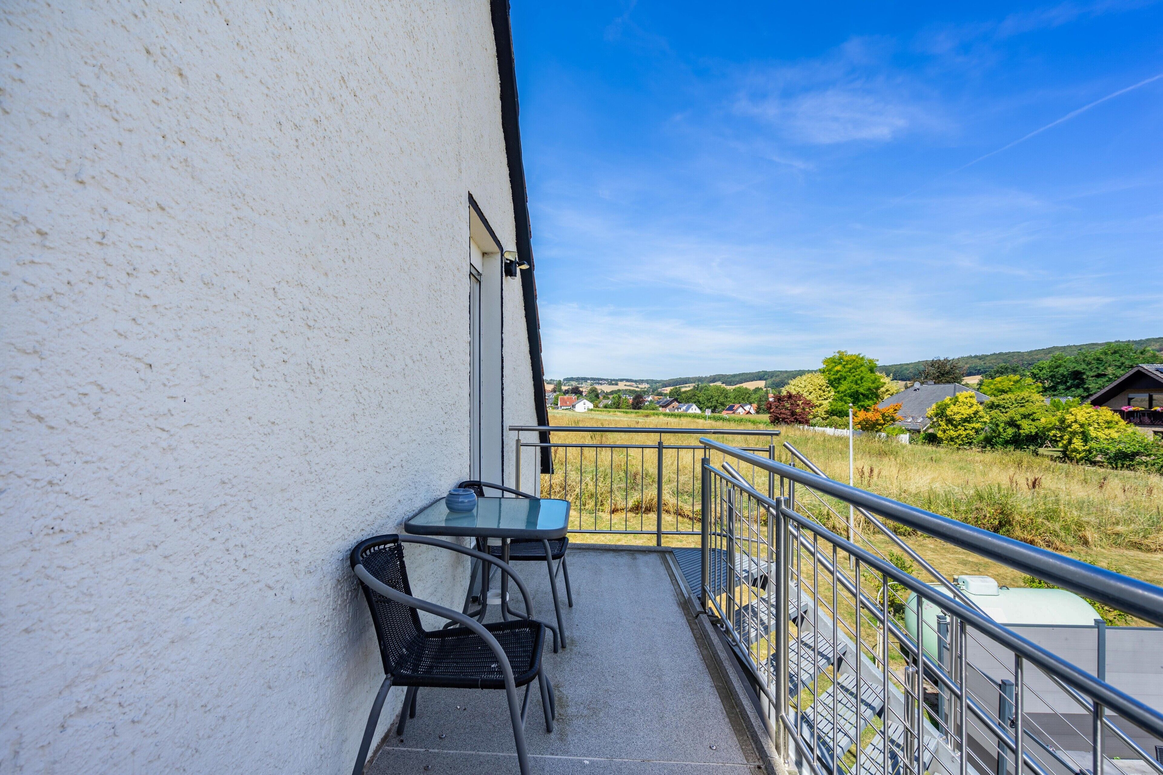 Ein Balkon mit Tisch und Stühlen, der auf eine ländliche Gegend mit Häusern und Feldern blickt.