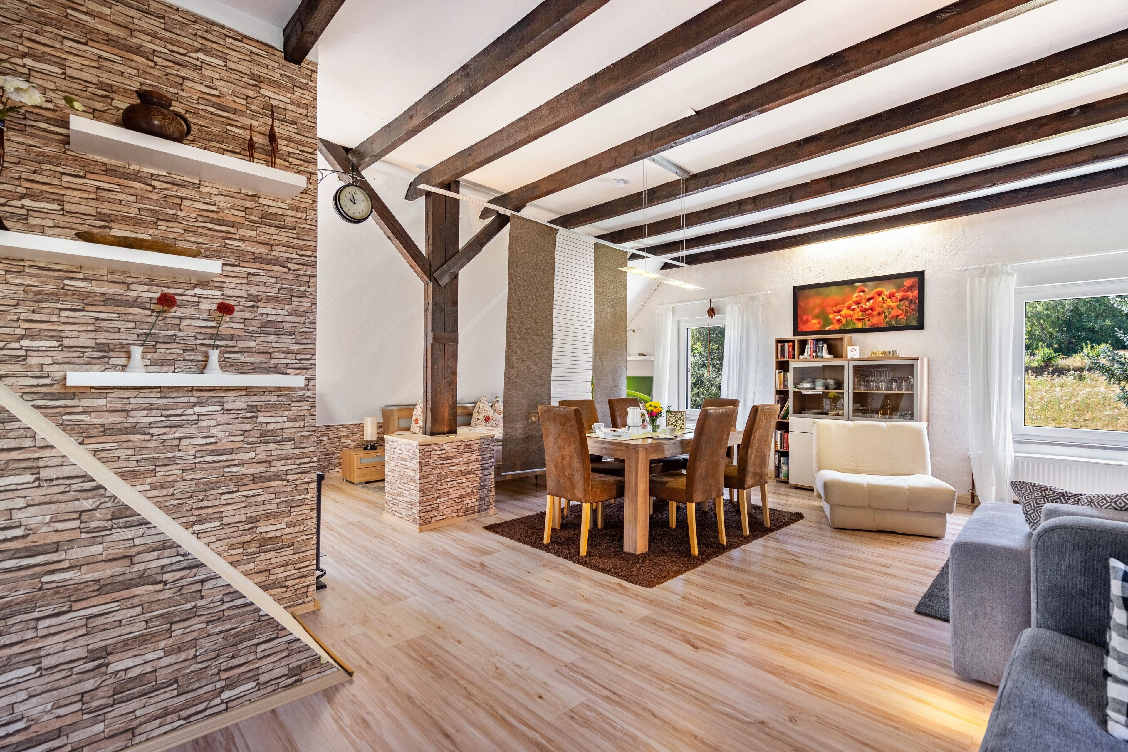 Ein modernes Wohnzimmer mit Steinwand, Holzbalken, einem Essbereich mit Tisch und Stühlen sowie einem Bücherregal.