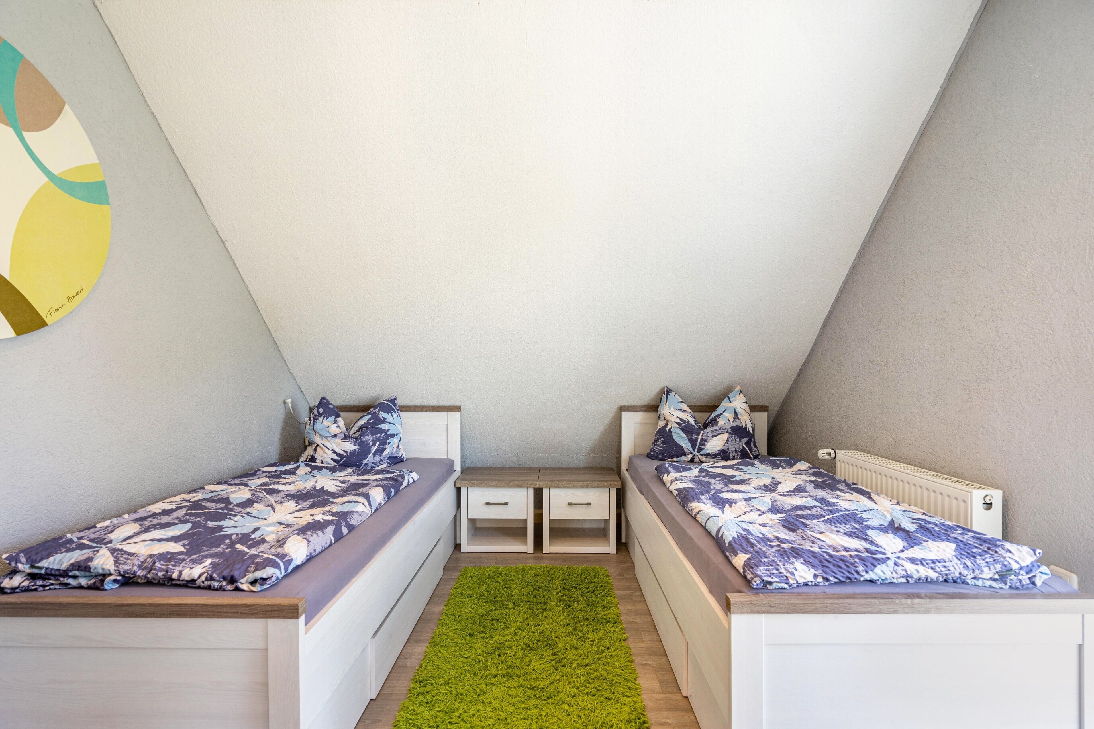Een kleine, driehoekige kamer met twee bedden, een groen tapijt en een witte muur met een gedeeltelijk abstract schilderij.