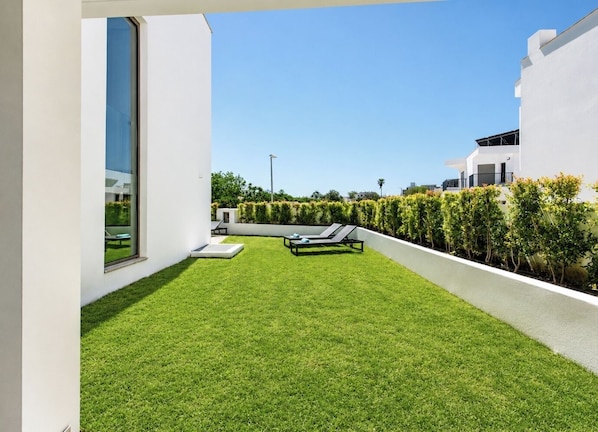 A modern outdoor space with a neatly trimmed lawn, two lounge chairs, and a white building with large windows.