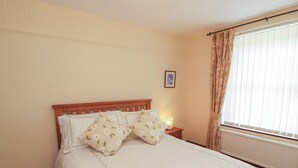 Een net opgemaakt bed met bloemenkussens, een bedlampje en een raam met gordijnen.